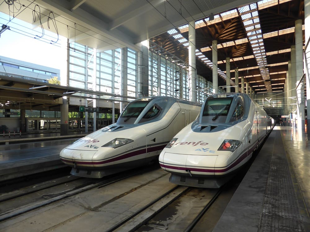 Two AVE high-speed trains side by side at a bustling Spanish train station.